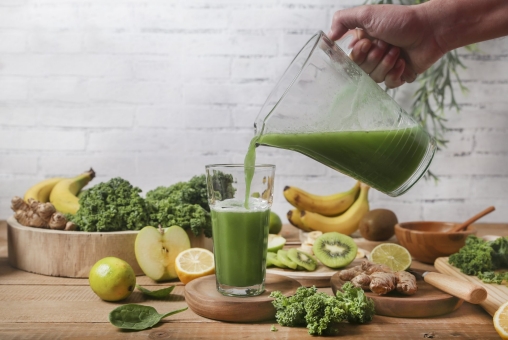 Es wird ein grüner Smoothie aus einer Glaskaraffe in ein Glas geschüttet. Im Hintergrund liegt viel Obst und Gemüse.