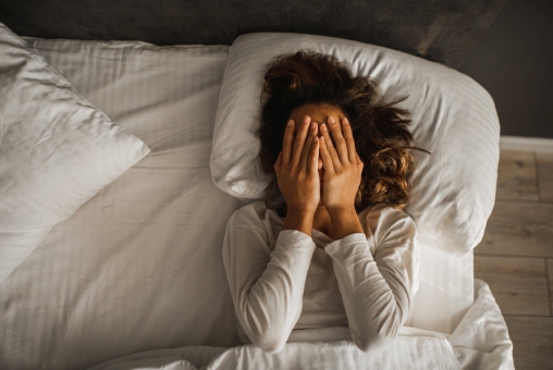 Eine Frau liegt im gestresst im Bett und schlägt die Hände vor ihrem Gesicht zusammen.