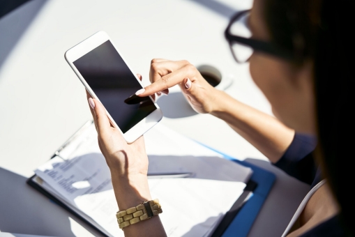 Eine junge Frau sitzt am Schreibtisch. Vor ihr liegen Dokumente in einem Klemmbrett und sie tippt auf ihrem Handy.