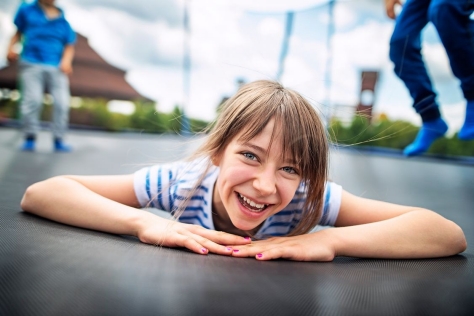 Ein junges Mädchen liegt auf dem Bauch und lächelt in die Kamera. Sie liegt auf einem Trampolin und im Hintergrund hüpfen weitere Kinder.