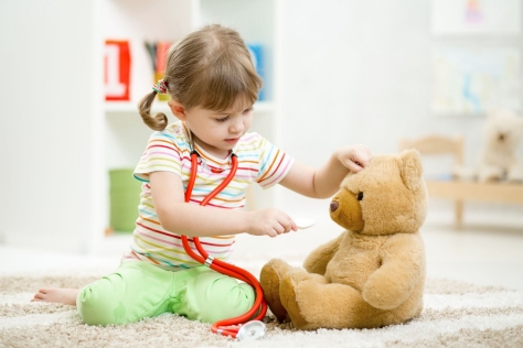Ein kleines Mädchen hat sich als Ärztin verkleidet und spielt mit einem großen Teddy-Bären.