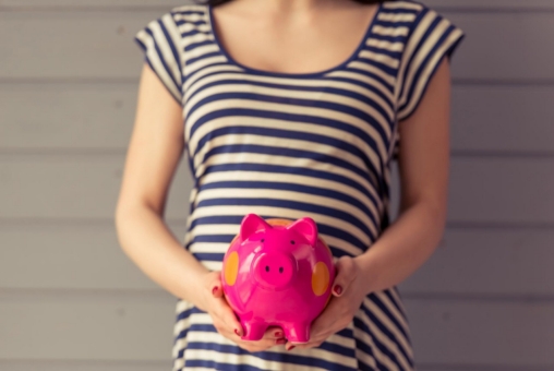 Eine schwangere Frau hält ein Sparschwein in der Hand vor ihren Bauch.