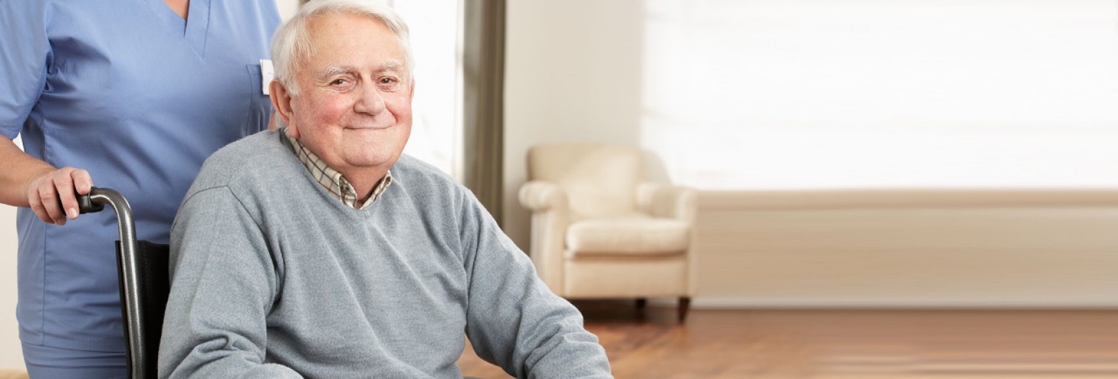 Ein Senior sitzt im Rollstuhl. Hinter ihm steht eine Pflegekraft und schiebt ihn.