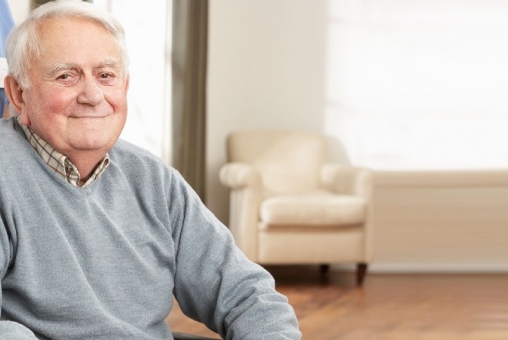 Ein Senior sitzt im Rollstuhl. Hinter ihm steht eine Pflegekraft und schiebt ihn.
