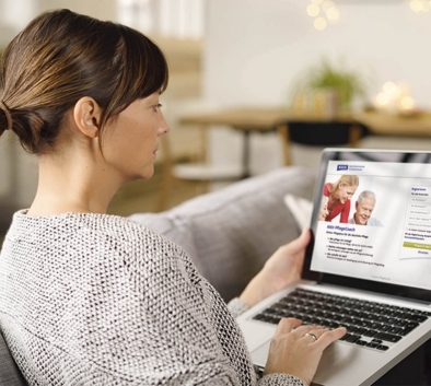 Eine Frau sitzt auf dem Sofa im Wohnzimmer und schaut auf ihren Laptop. Sie hat die Seite vom Online Pflegecoach der KKH geöffnet.