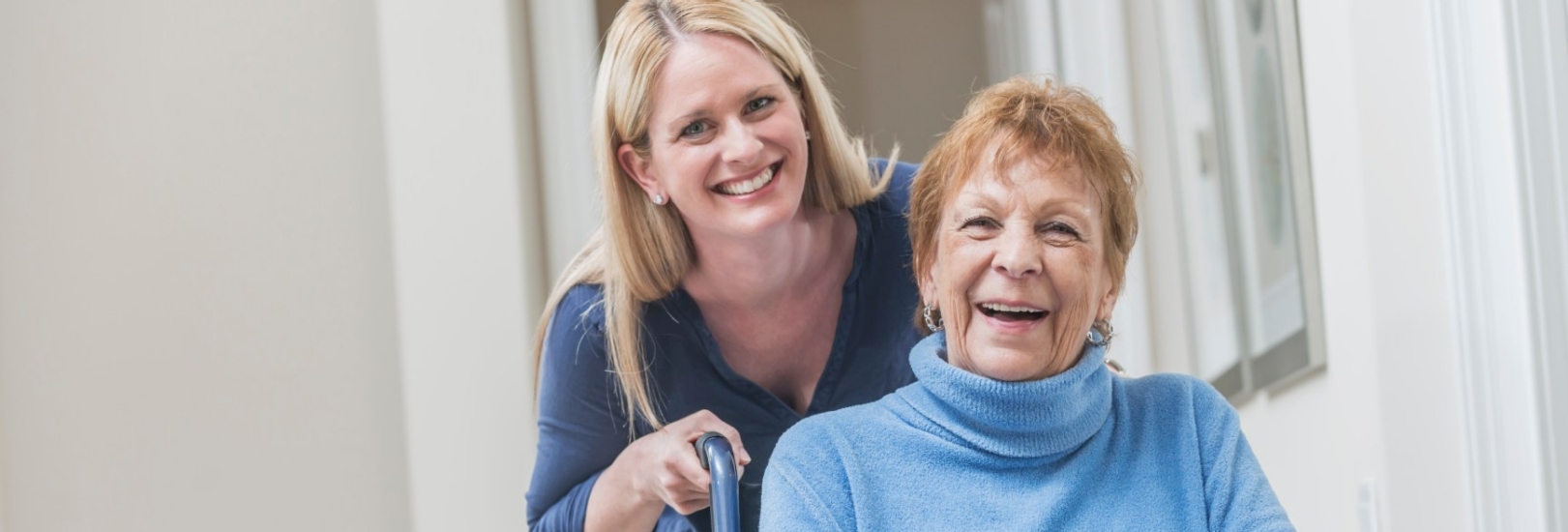 Eine jüngere Frau schiebt eine lächelnde Seniorin im Rollstuhl.