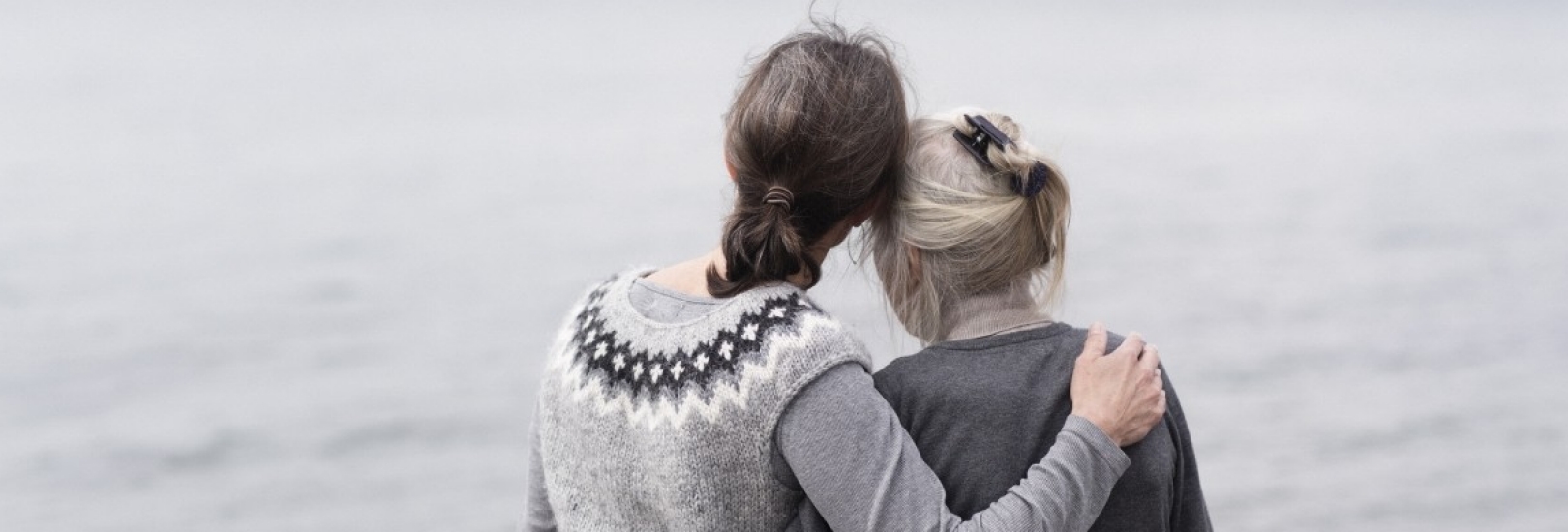 Eine Frau und eine Seniorin blicken auf das vor ihnen liegende Wasser und halten sich in den Armen.