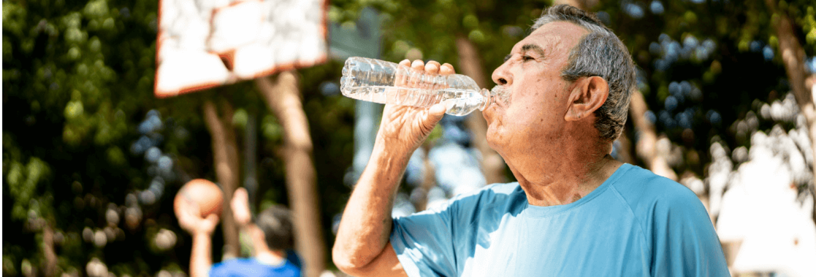 Ein älterer Mann trinkt aus einer Wasserflasche. Im Hintergrund ist ein Basketballfeld.