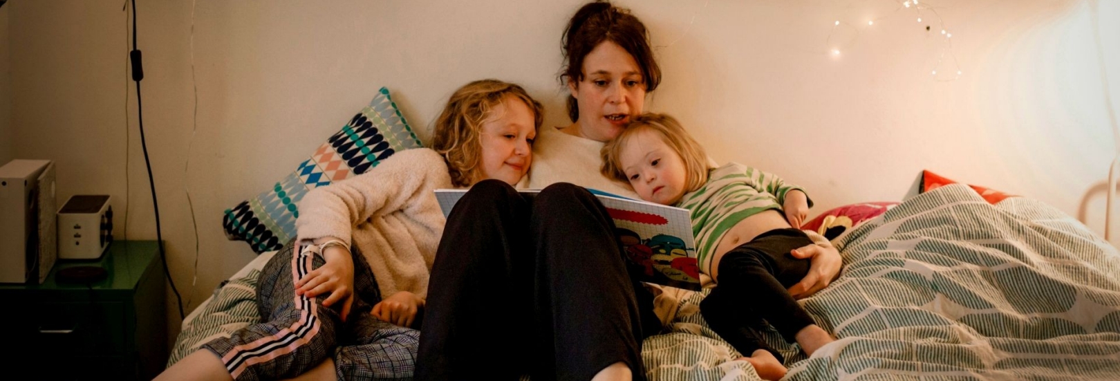 Frau liegt mit ihren beiden Kindern im Bett und liest aus einem Buch vor.