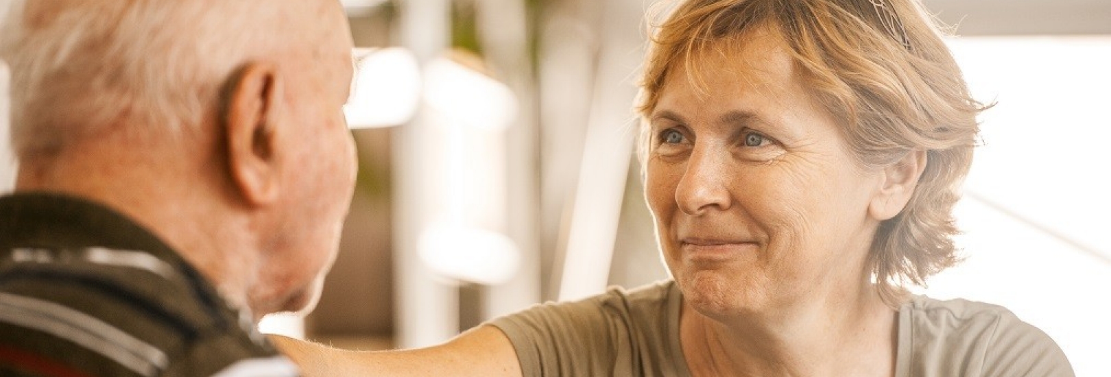 Eine Tochter besucht ihren älteren Vater in Pflegeeinrichtung. Sie lächelt.