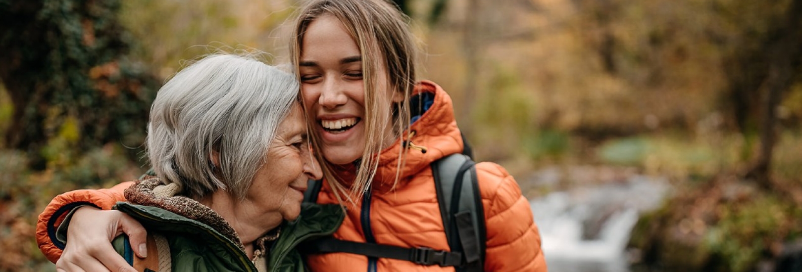 Eine Seniorin und eine Enkelin sind auf einem Waldspaziergang zu sehen. Beide umarmen sich glücklich.