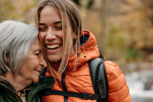 Eine Seniorin und eine Enkelin sind auf einem Waldspaziergang zu sehen. Beide umarmen sich glücklich.