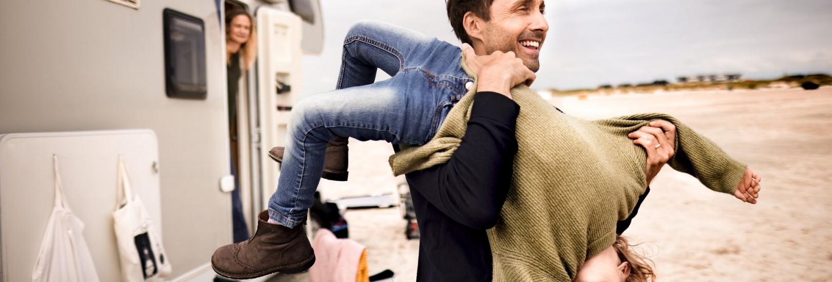 Ein junger Vater hält sein Kind Kopfüber in den Armen. Sie machen eine Wohnwagentour und haben am Strand geparkt.
