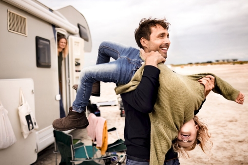 Ein junger Vater hält sein Kind Kopfüber in den Armen. Sie machen eine Wohnwagentour und haben am Strand geparkt.