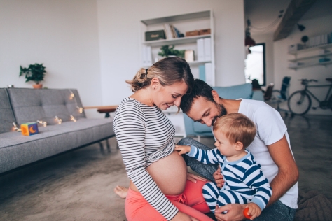 Eine junge Familie sitzt im Wohnzimmer. Die Mutter hat einen Babybauch und lässt ihr Kleinkind darüber streichen.