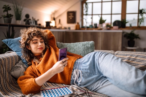 Lächelnde junge Frau entspannt sich auf dem Sofa in ihrem Wohnzimmer, während sie ihr mobiles Smartphone für die myCoach-App nutzt